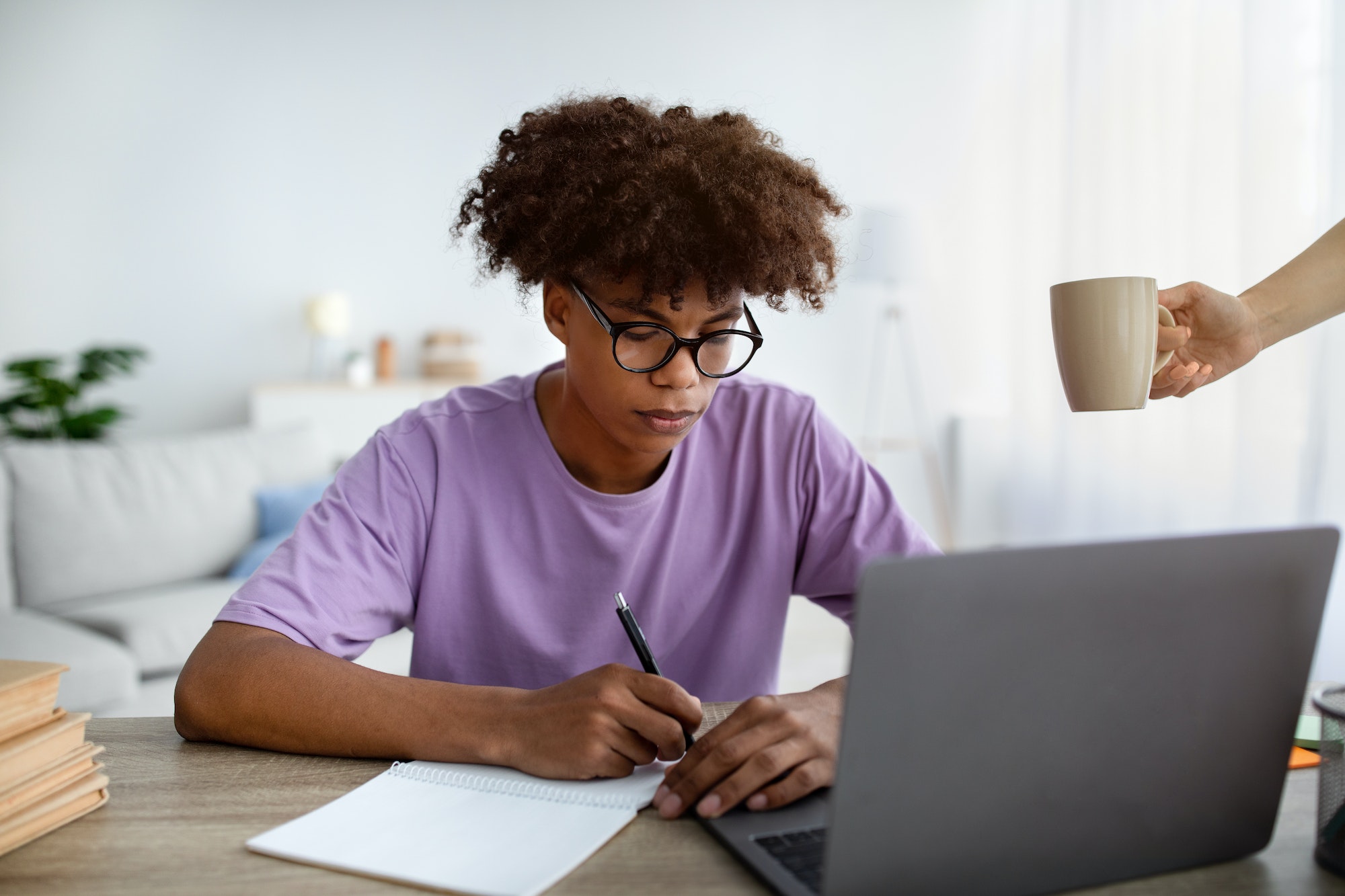 Serious black teen guy writing in notebook during webinar or lesson on laptop at home